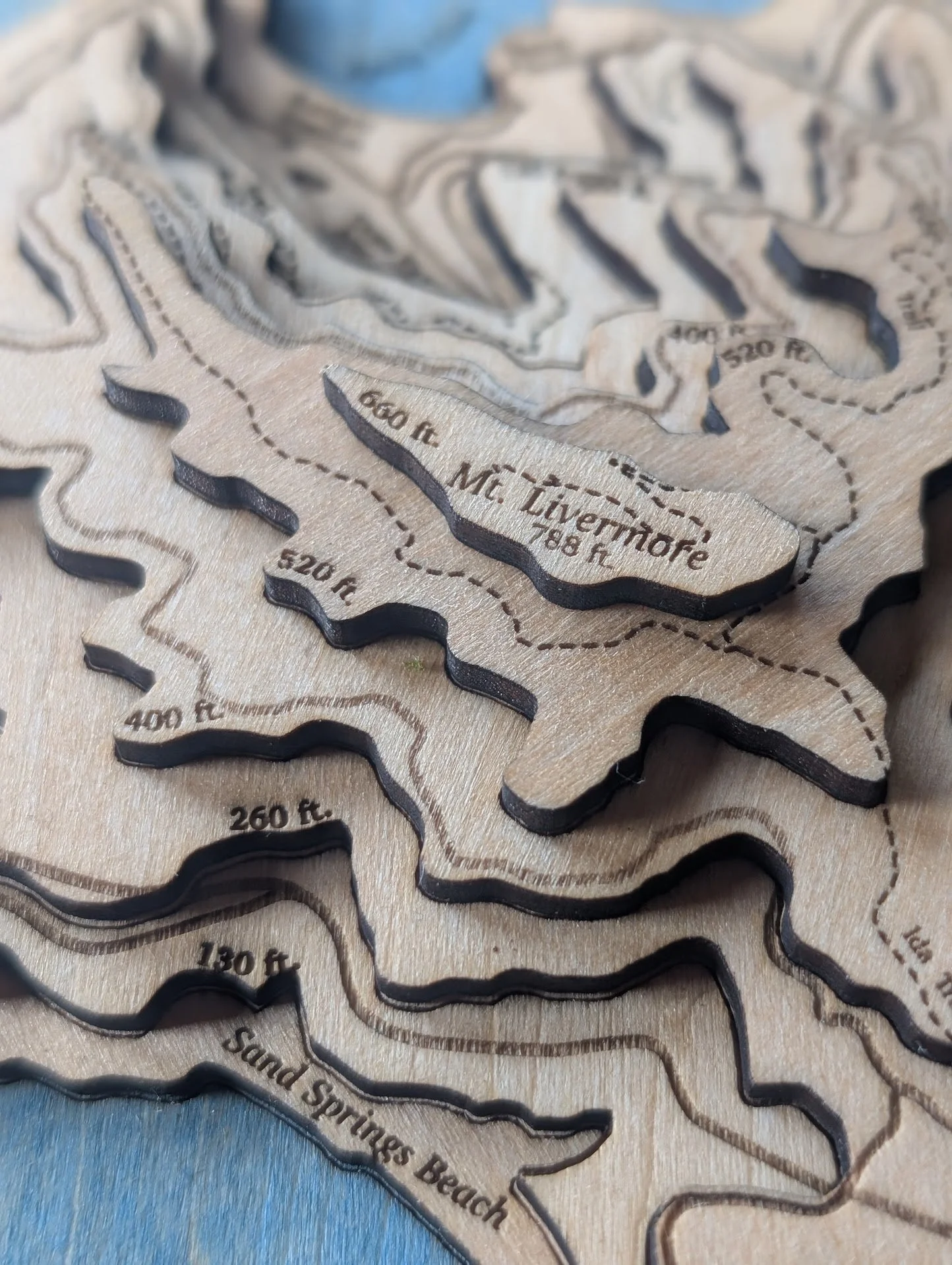 Macro detail of the Angel Island topography showing elevation labels etched into each layer — 130ft, 260ft, 400ft, 520ft, 600ft — climbing toward Mt. Livermore at 788ft. Engraved dashed lines indicate hiking trails, with Sand Springs Beach labeled at the shore.