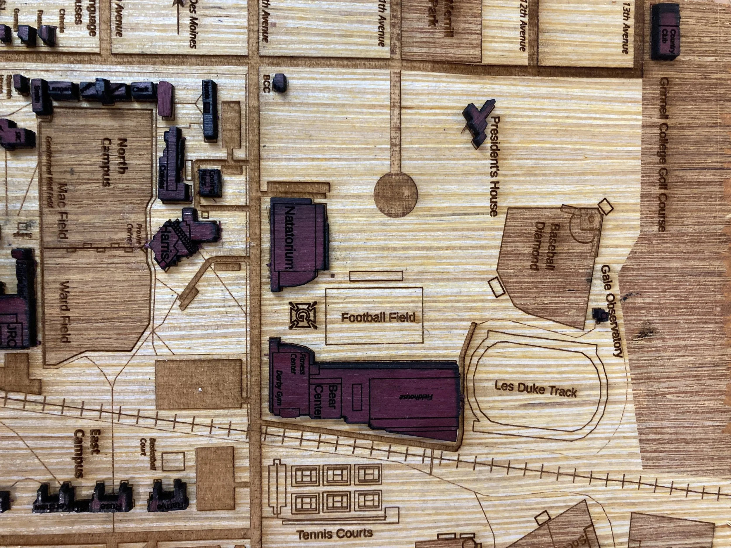 Detail of the Grinnell map's athletic complex showing Les Duke Track, the football field, tennis courts, and surrounding campus buildings in purple wood. Street names like 8th Avenue are engraved into the base, along with the railroad tracks that bisect the town.