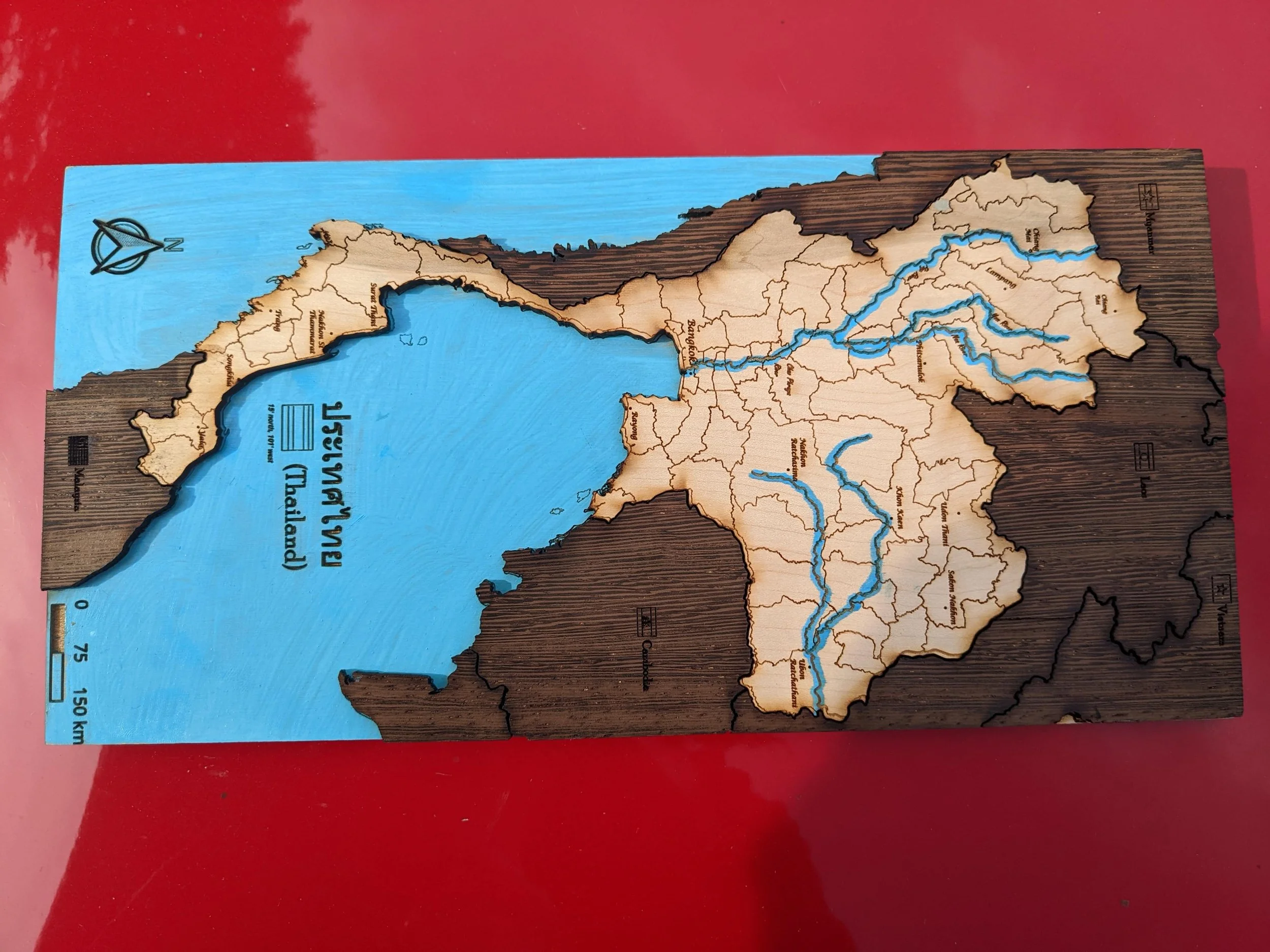 The Thailand map laid flat on a red surface, viewed from above. This angle emphasizes the country's geography — the narrow southern peninsula reaching toward Malaysia, the broad northeastern Isan region bordering Laos, and the branching blue river systems flowing toward the gulf.