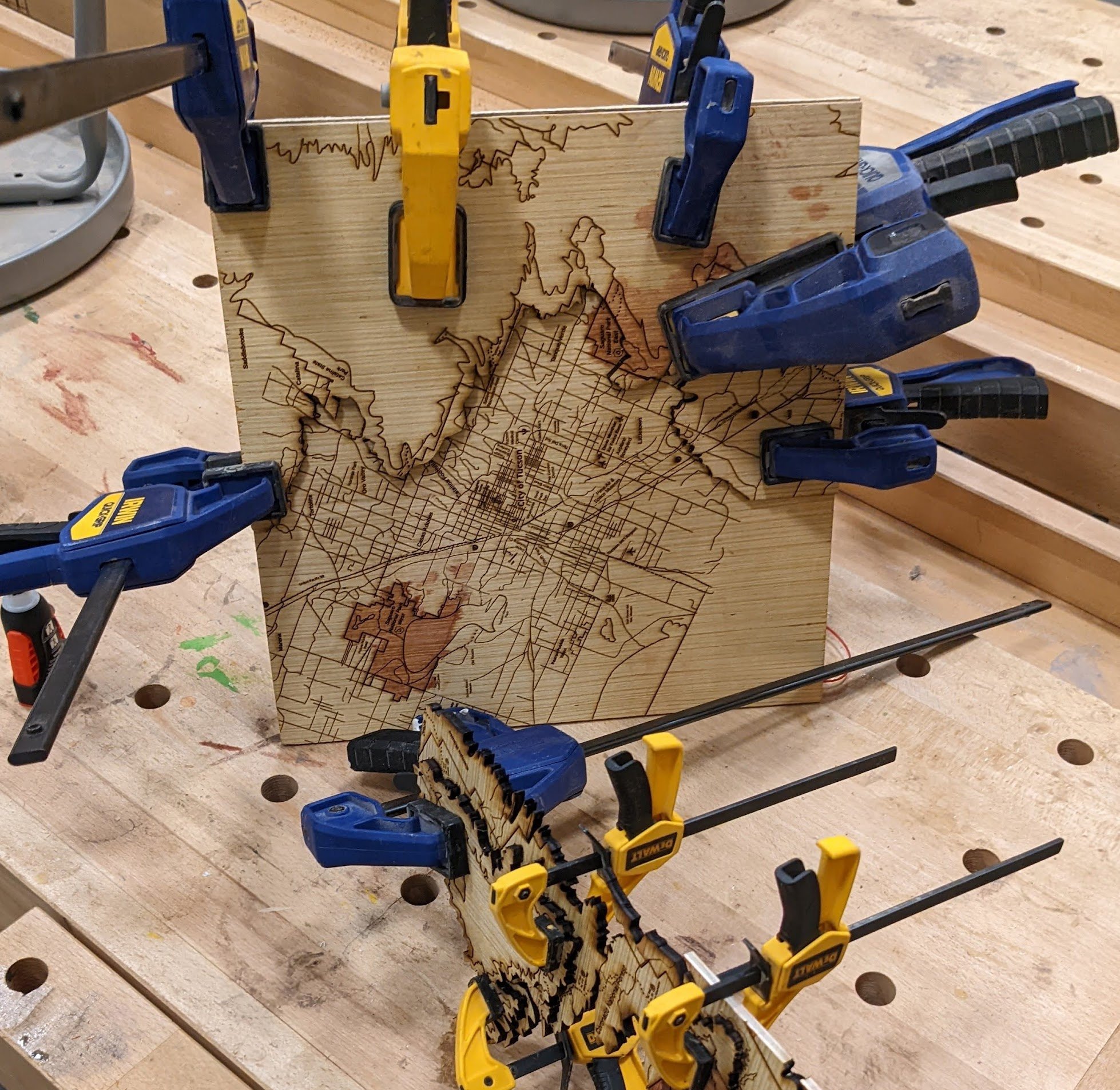 Two sections of a laser-cut wooden topographic map of Tucson, Arizona held in bar clamps during assembly. The larger 12x12 inch square piece shows the city's street grid overlaid with contour lines, while the smaller irregular piece displays dramatic layered terrain with elevation changes visible through the cut plywood laminations.